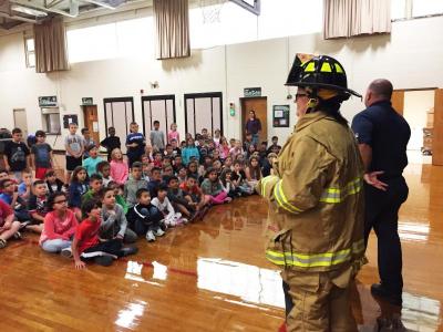 Students gawk and giggle as third grade teacher Amanda Depin tries on a firefighter’s uniform.