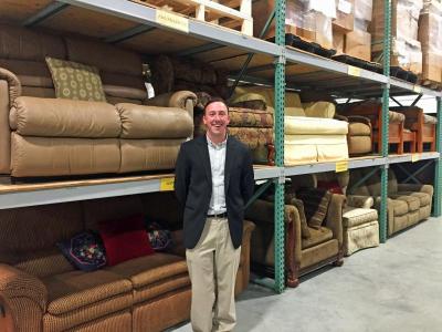 Josh Smith, director of the organization’s Dartmouth facility, stands in the Reed Road warehouse in front of shelves of donated furniture.
